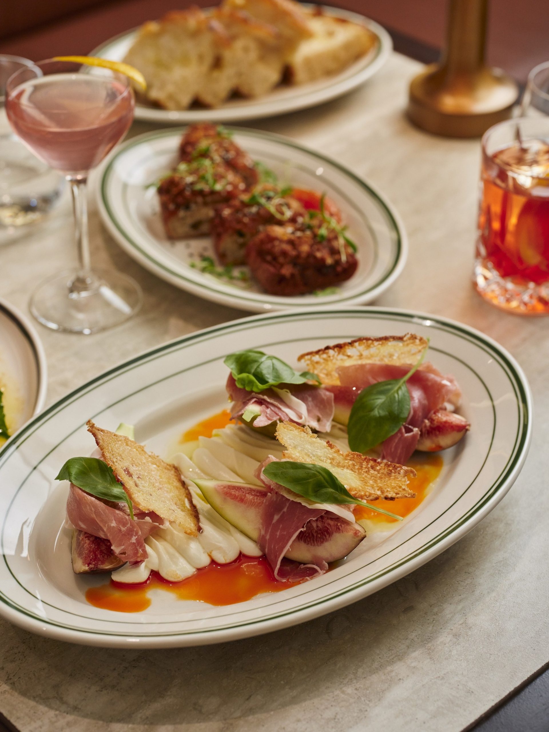 Prosciutto and fig antipasti plate with fennel, crostini, and basil, alongside cocktails and meatballs.