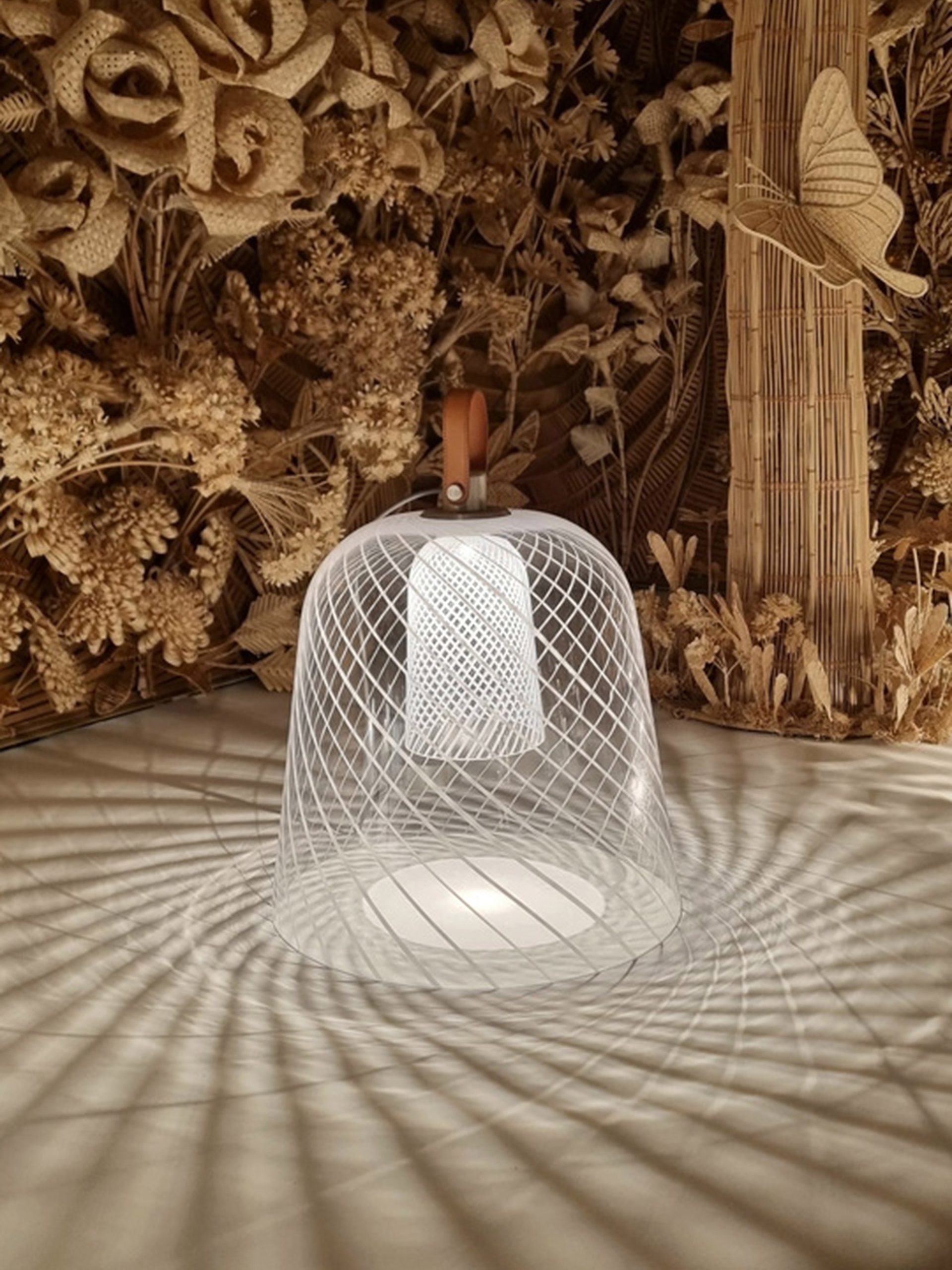 White wire mesh bell lamp with wooden handle casting lattice shadows before a dried botanical wall installation.