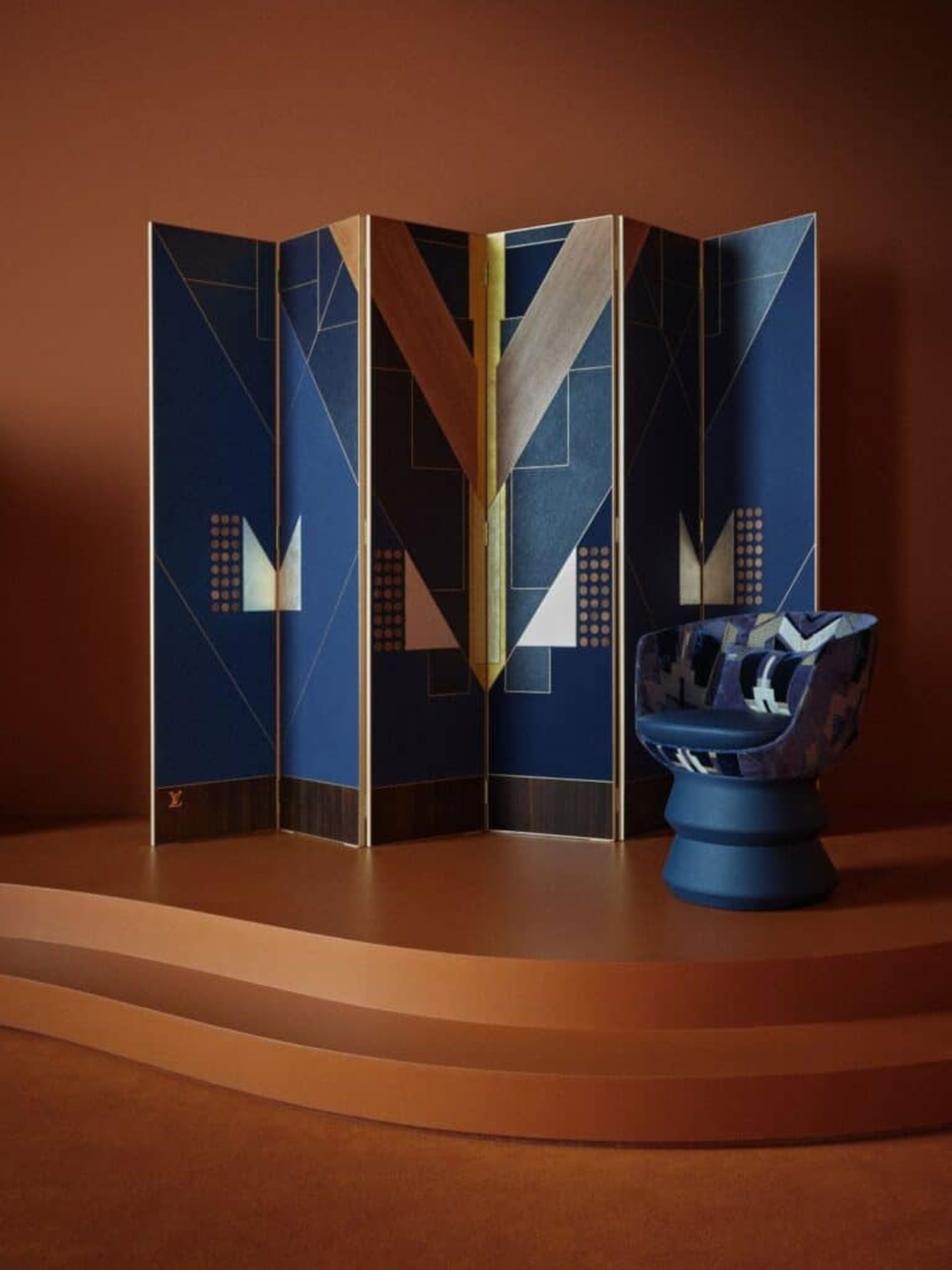 Louis Vuitton navy blue geometric folding screen with gold trim beside a sculptural armchair on a terracotta platform.