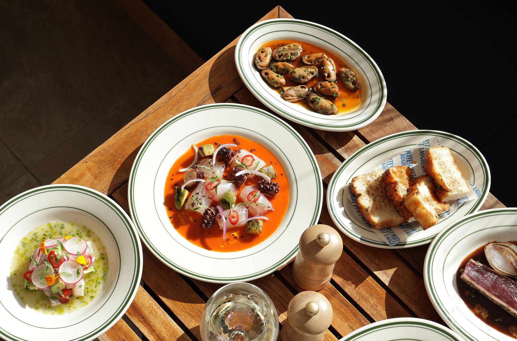 Overhead view of five Peruvian-inspired seafood dishes on green-rimmed white plates on a wooden table.