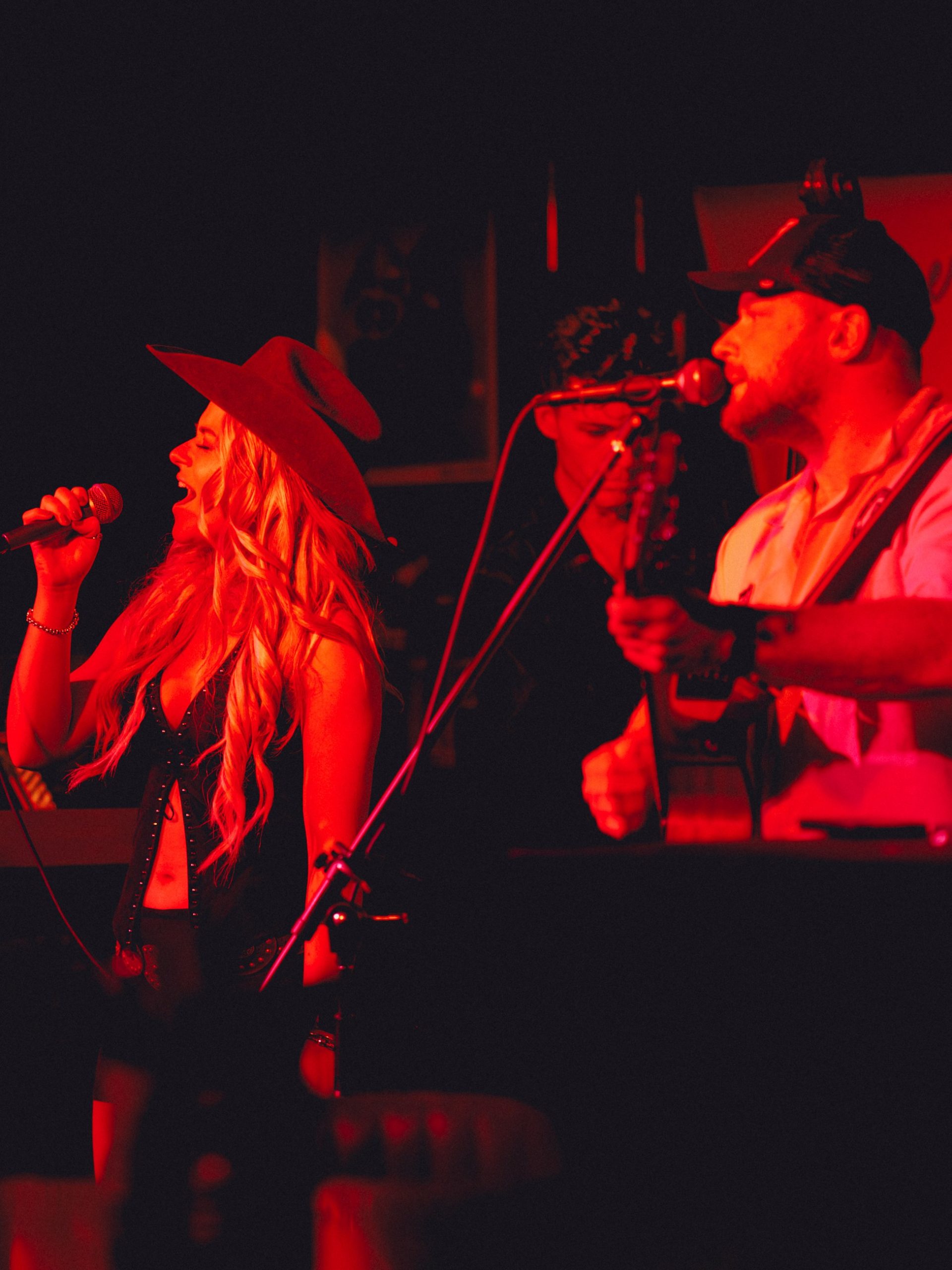 Blonde female singer in cowboy hat performs on a red-lit stage with band members.