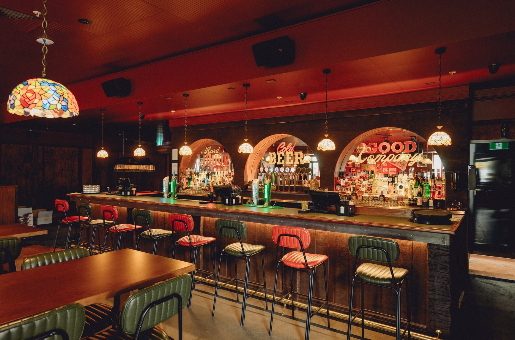 Warmly lit pub interior with red and green bar stools, arched neon signs, and Tiffany pendant lights.