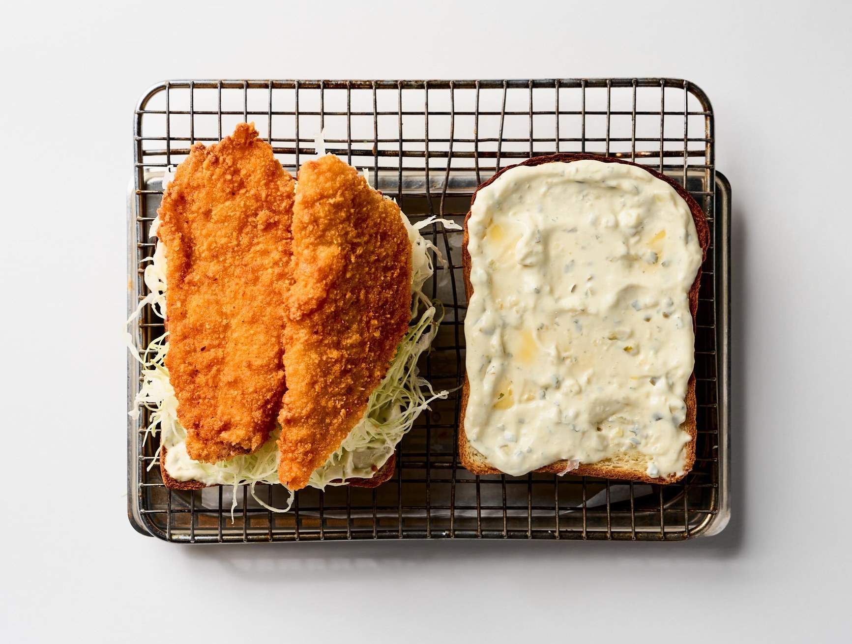 Open crumbed fish sandwich with shredded cabbage and tartare sauce on a wire rack.