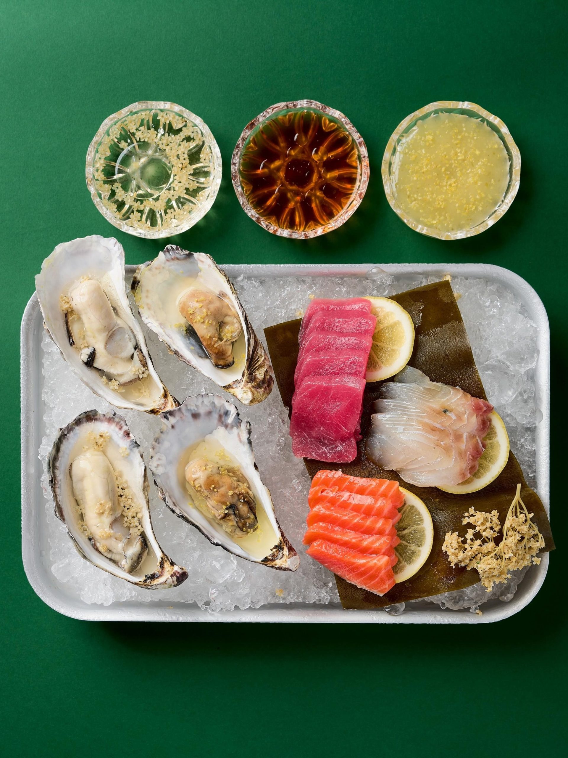 Overhead flat-lay of oysters and sashimi on crushed ice with three condiment bowls on green background.