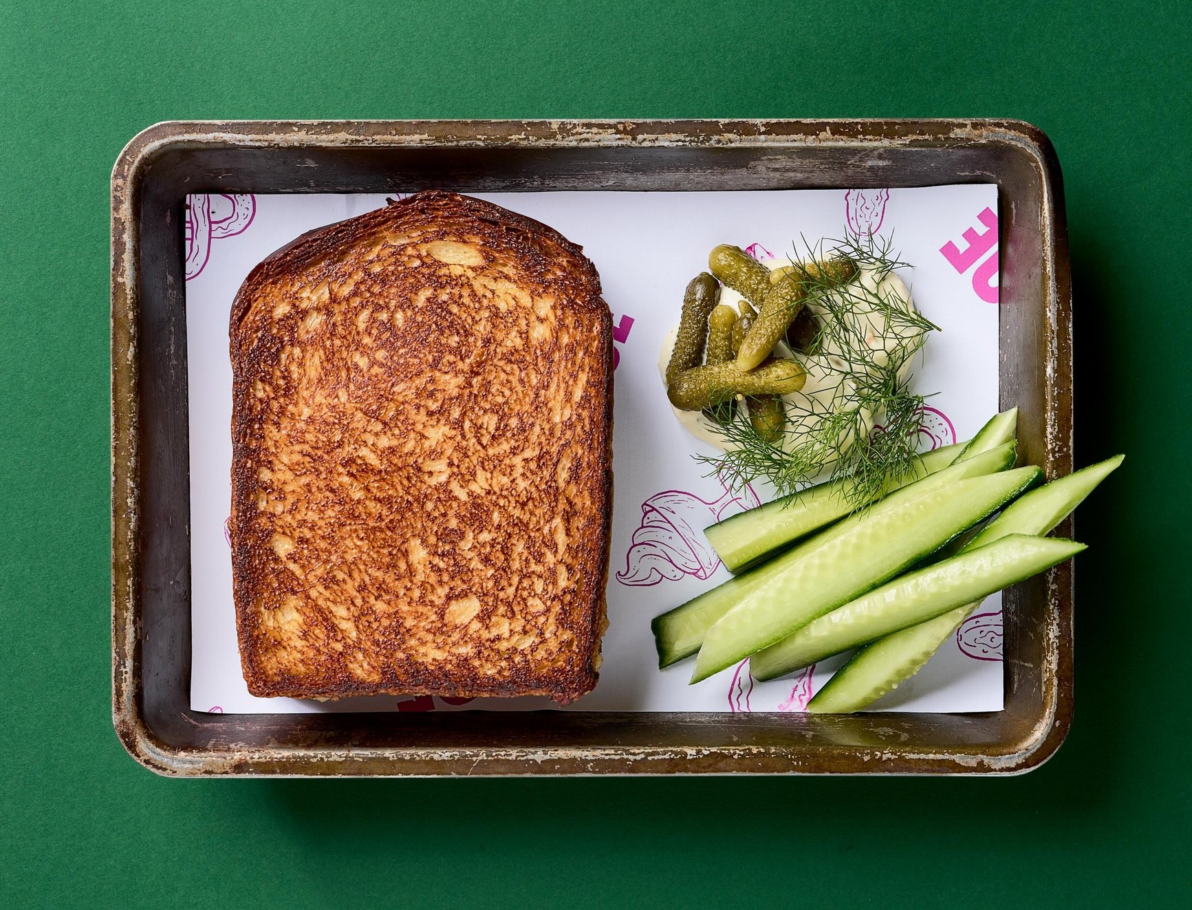 Overhead view of a toasted sandwich on a metal tray with cucumber, cornichons, and dill.