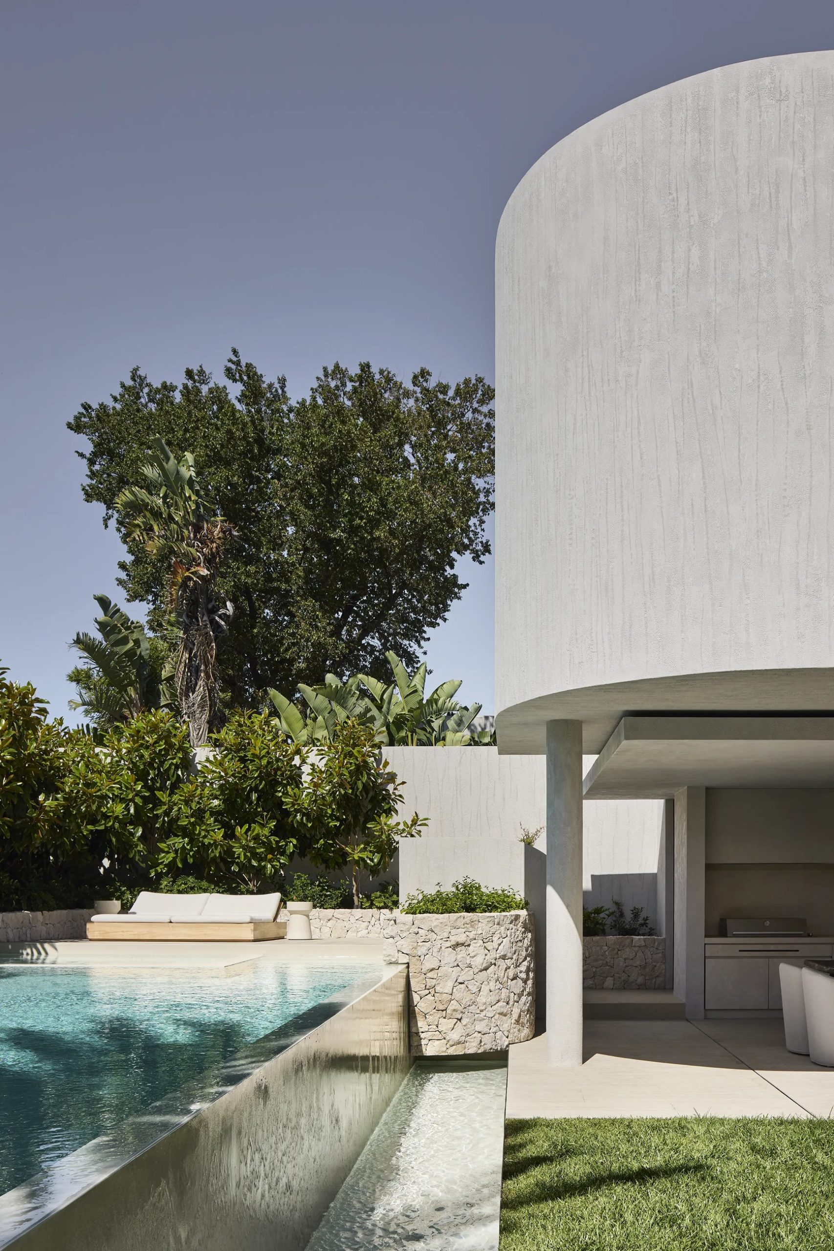Curved concrete tower beside infinity pool with outdoor kitchen pavilion and lush subtropical garden.