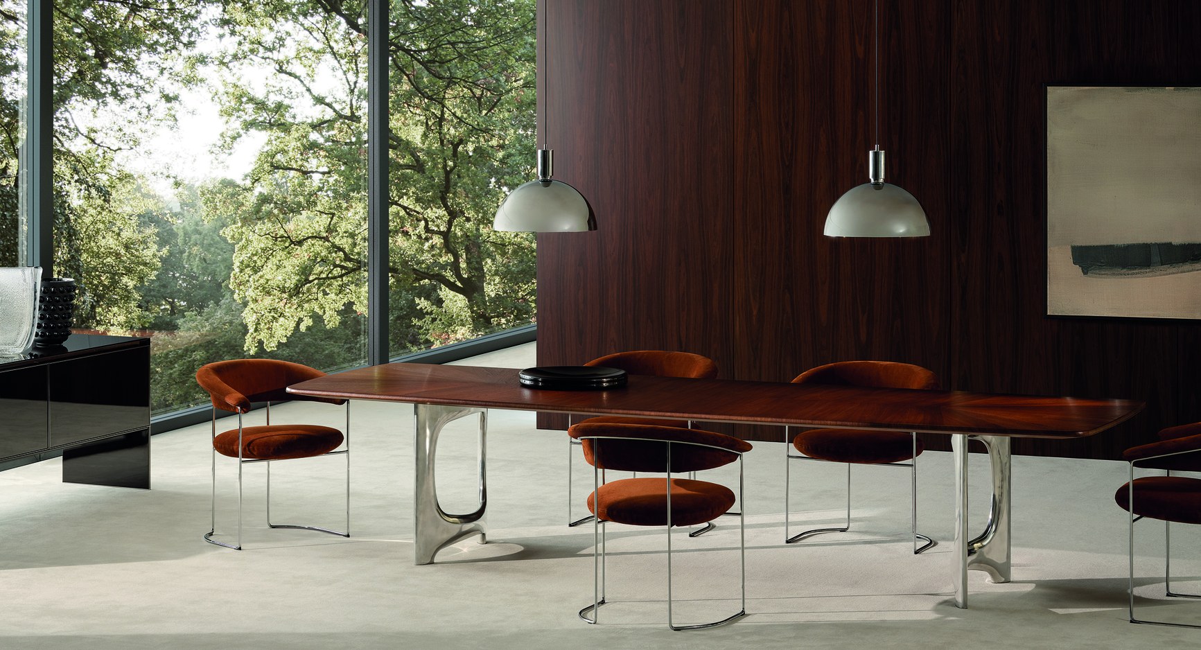 Rosewood dining table with chrome legs and rust velvet chairs in a modern room with forest views.