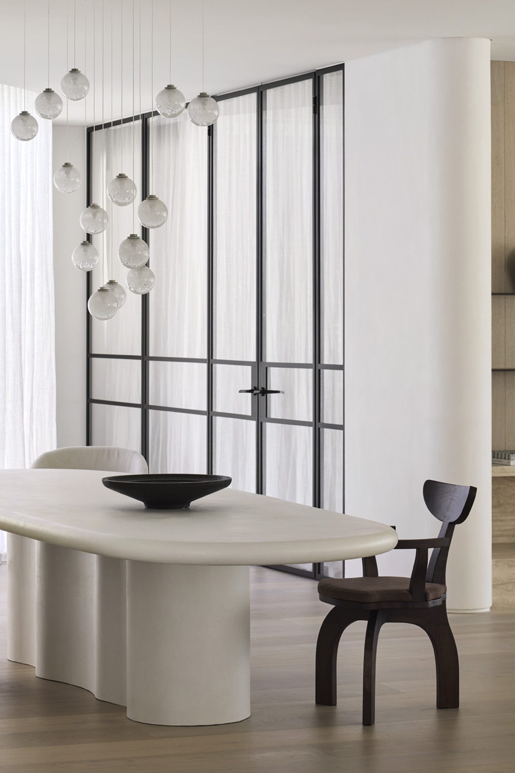 Minimalist dining room with cream sculptural table, globe pendant lights, and oak shelving with ceramic vessels.