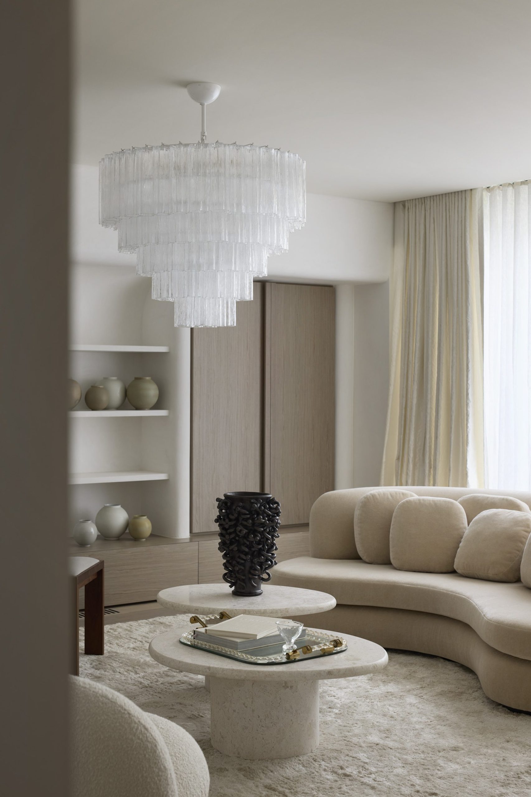 Luxury neutral living room with curved cream sofa, travertine coffee tables, and tiered glass chandelier.