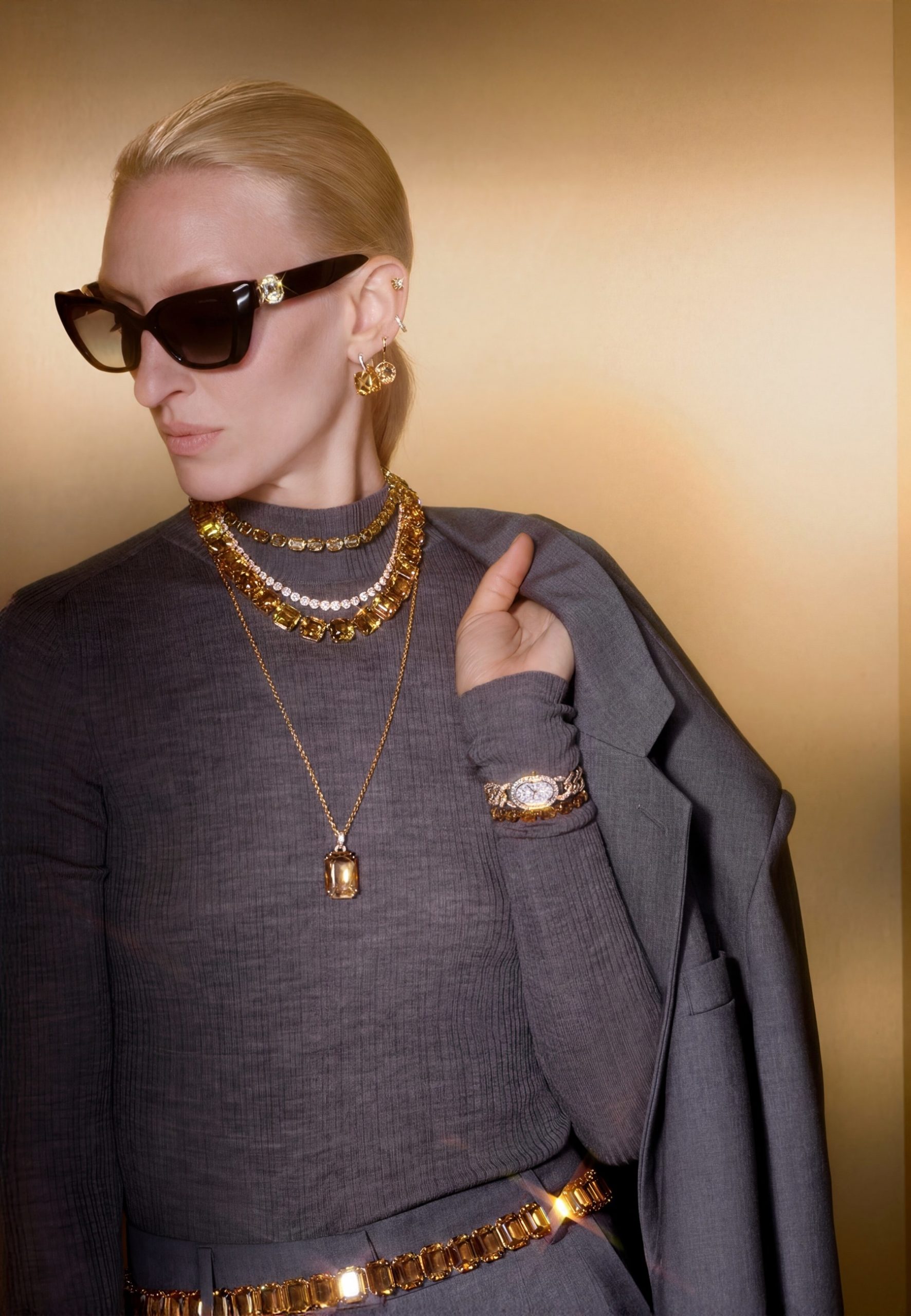 Blonde model in grey turtleneck layered with gold citrine jewellery, sunglasses, and chain belt in golden elevator.