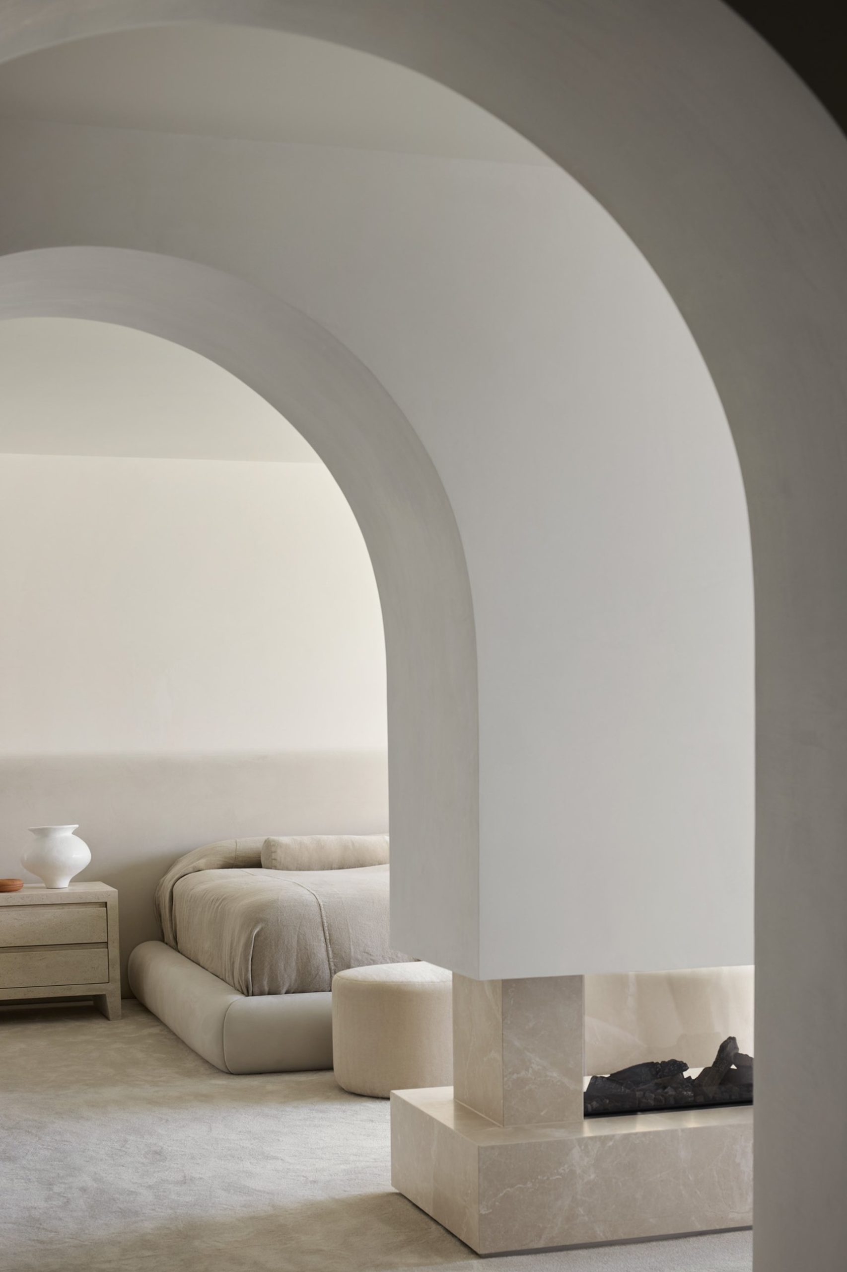 Minimalist interior with layered arches, travertine fireplace, and neutral upholstered bed beyond.