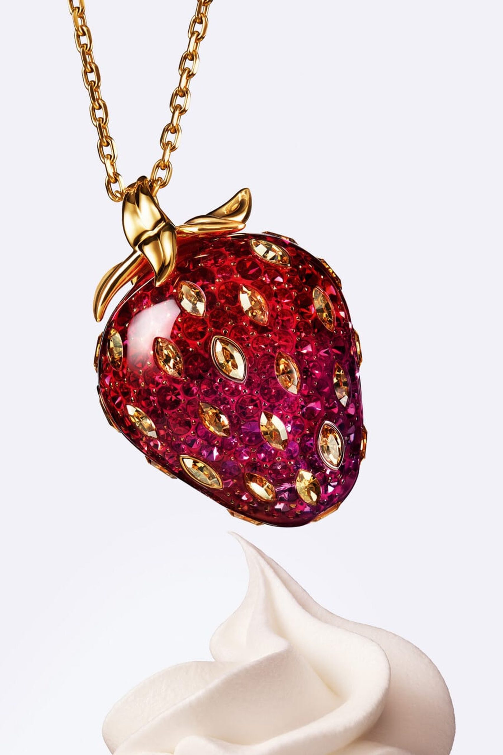 Gold-tone Swarovski strawberry pendant encrusted in red and amber crystals hovering above whipped cream.