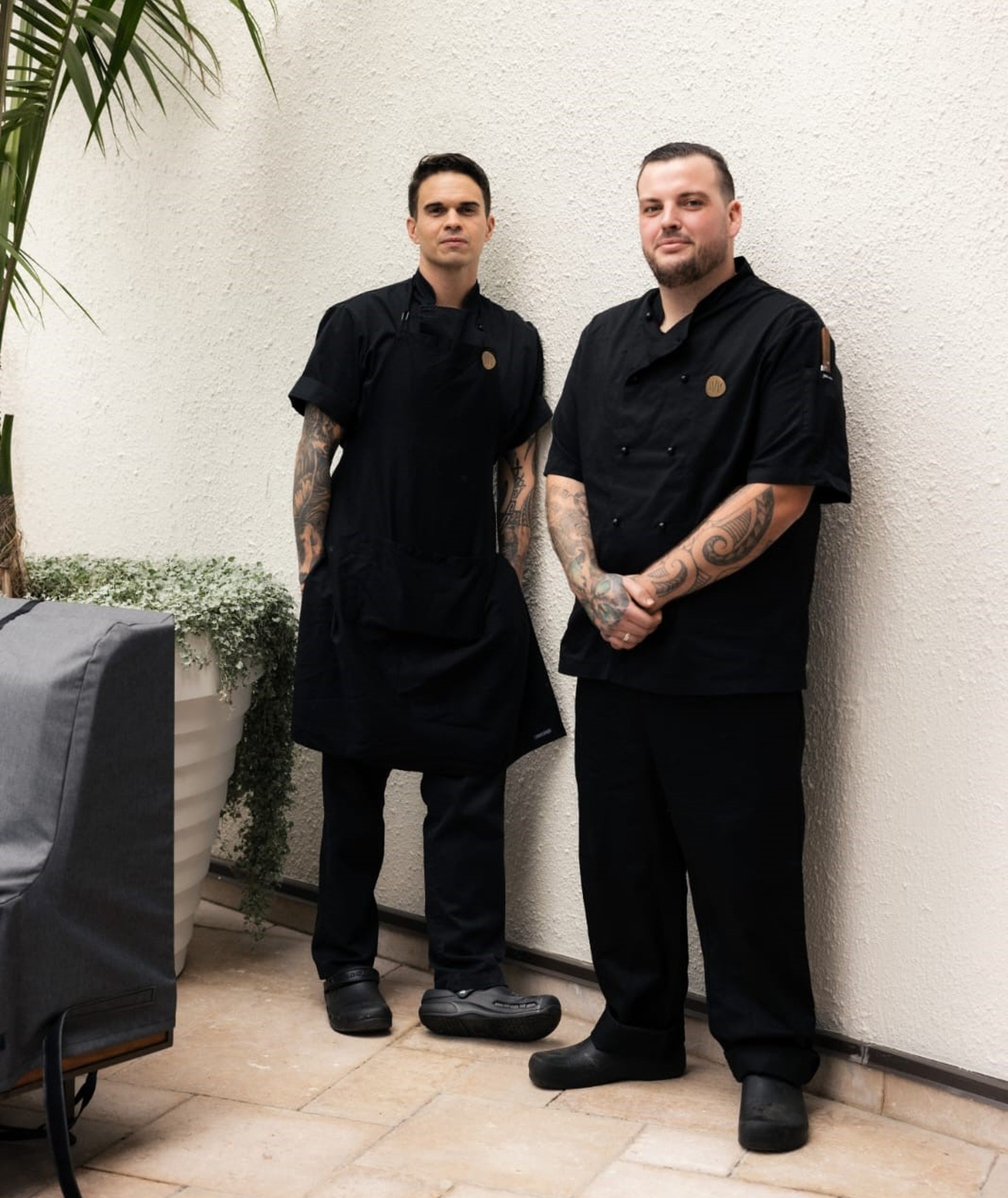 Two tattooed chefs in black uniforms standing against a white wall in an outdoor courtyard.
