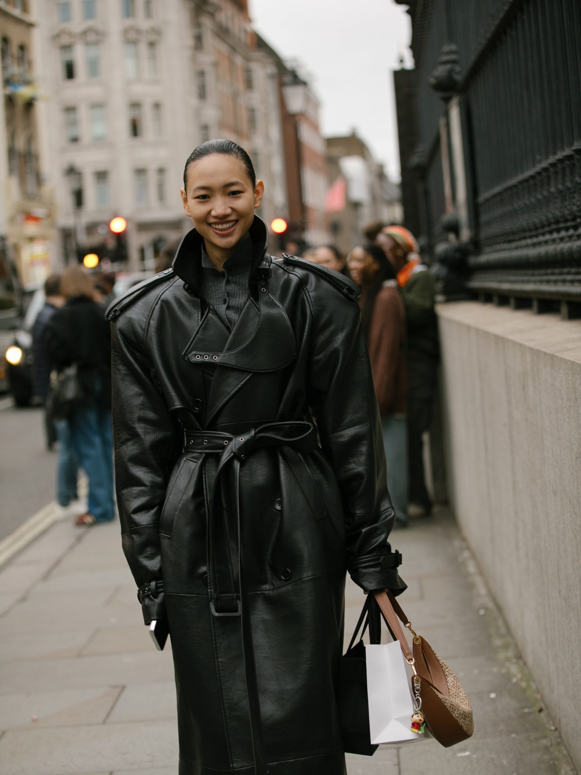 Woman in oversized black leather belted trench coat smiling on a London street.