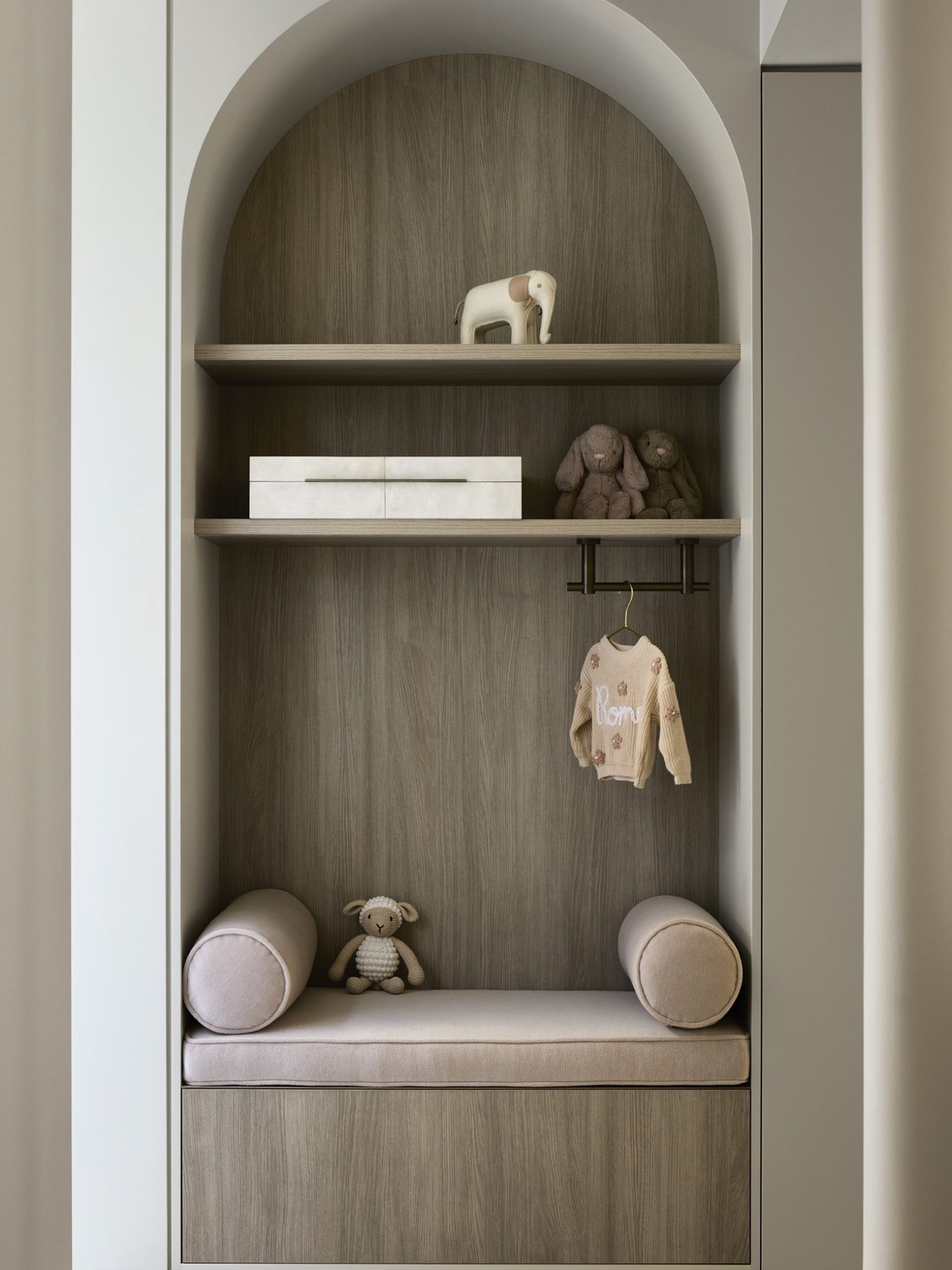 Arched timber built-in niche with blush bench seat, soft toys, and personalised children's knitwear.