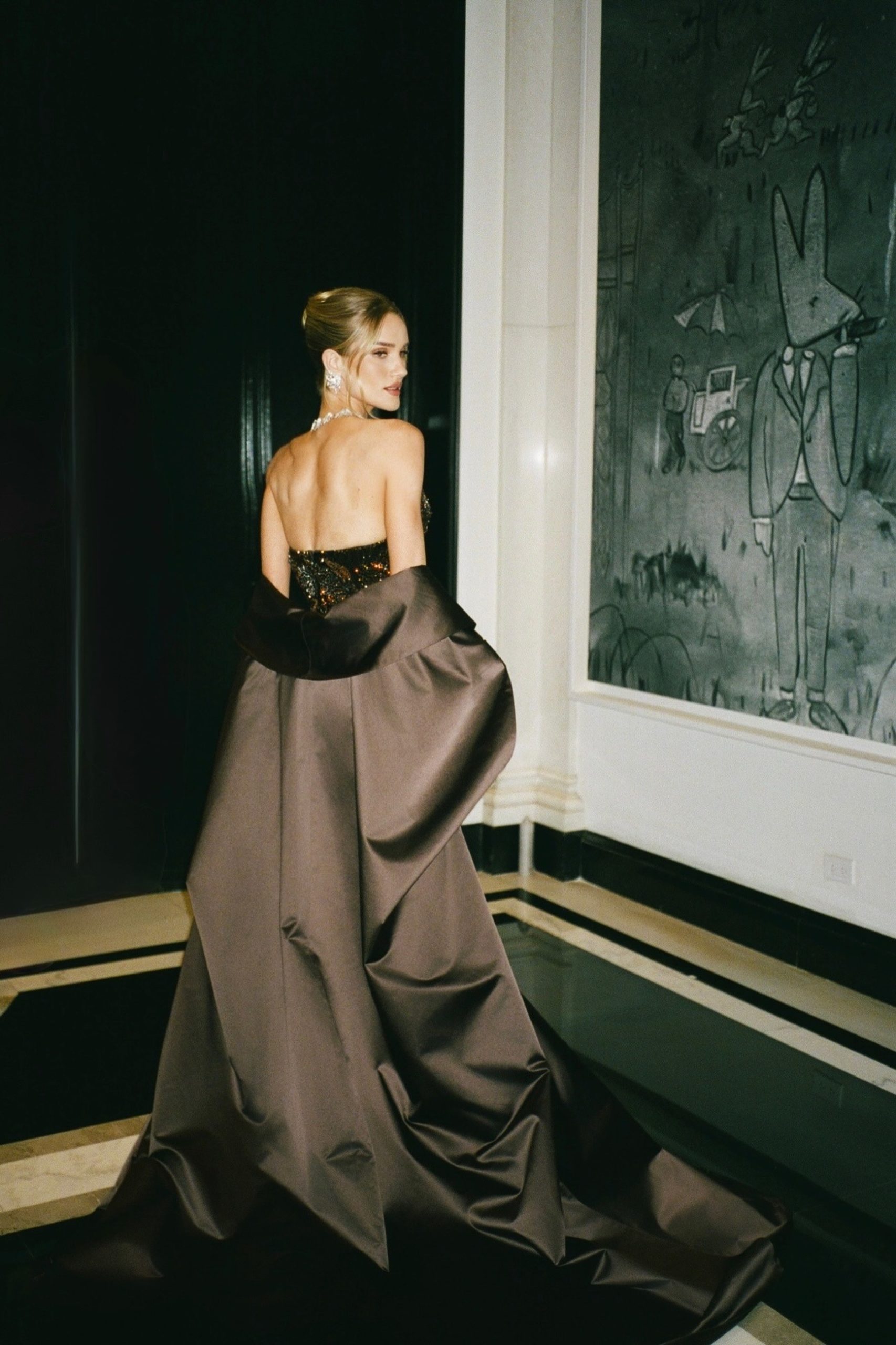Woman in dramatic dark satin strapless ballgown with train photographed from behind in luxury interior.