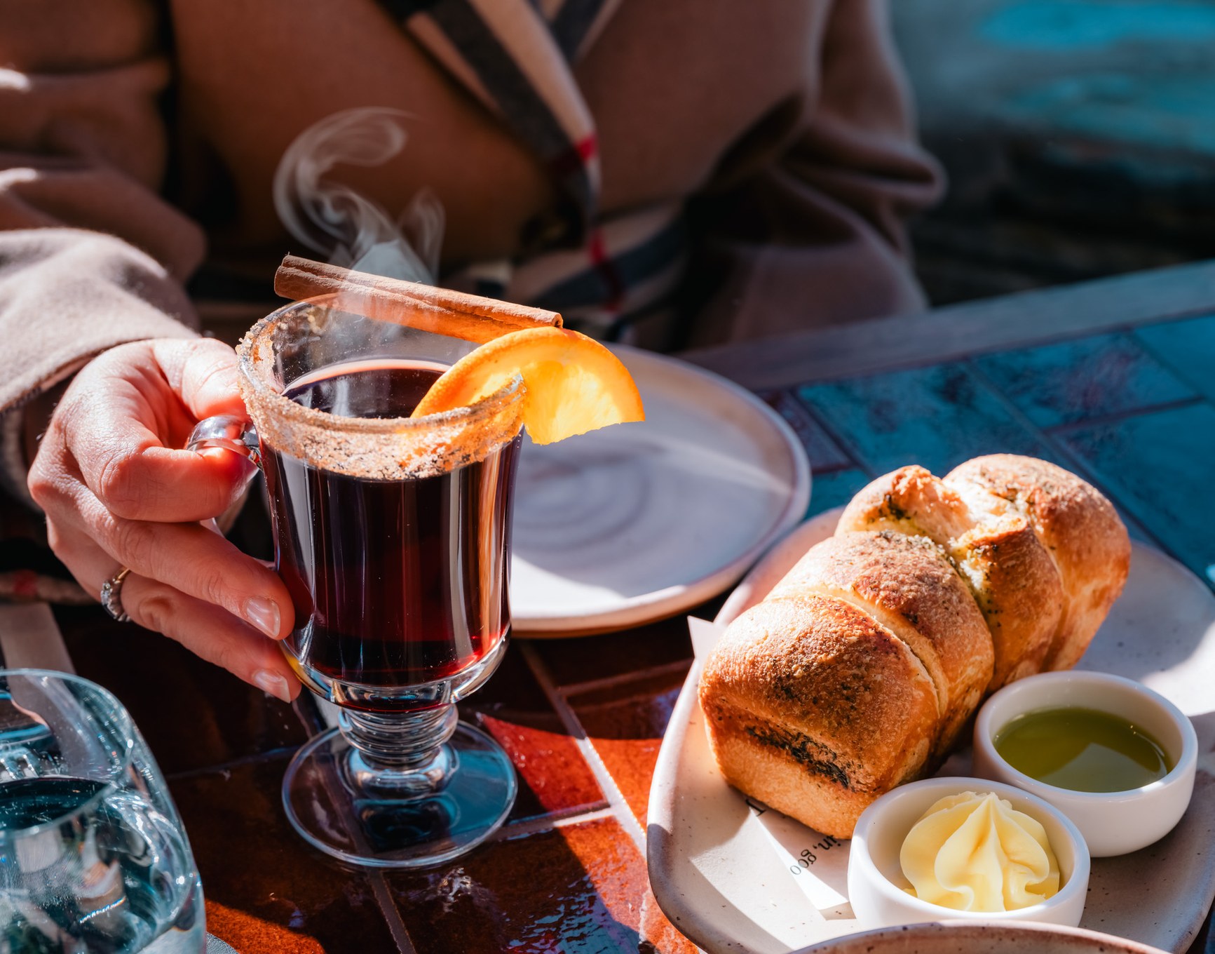 Hand holding a steaming mulled wine beside pull-apart bread rolls with butter and herb oil.