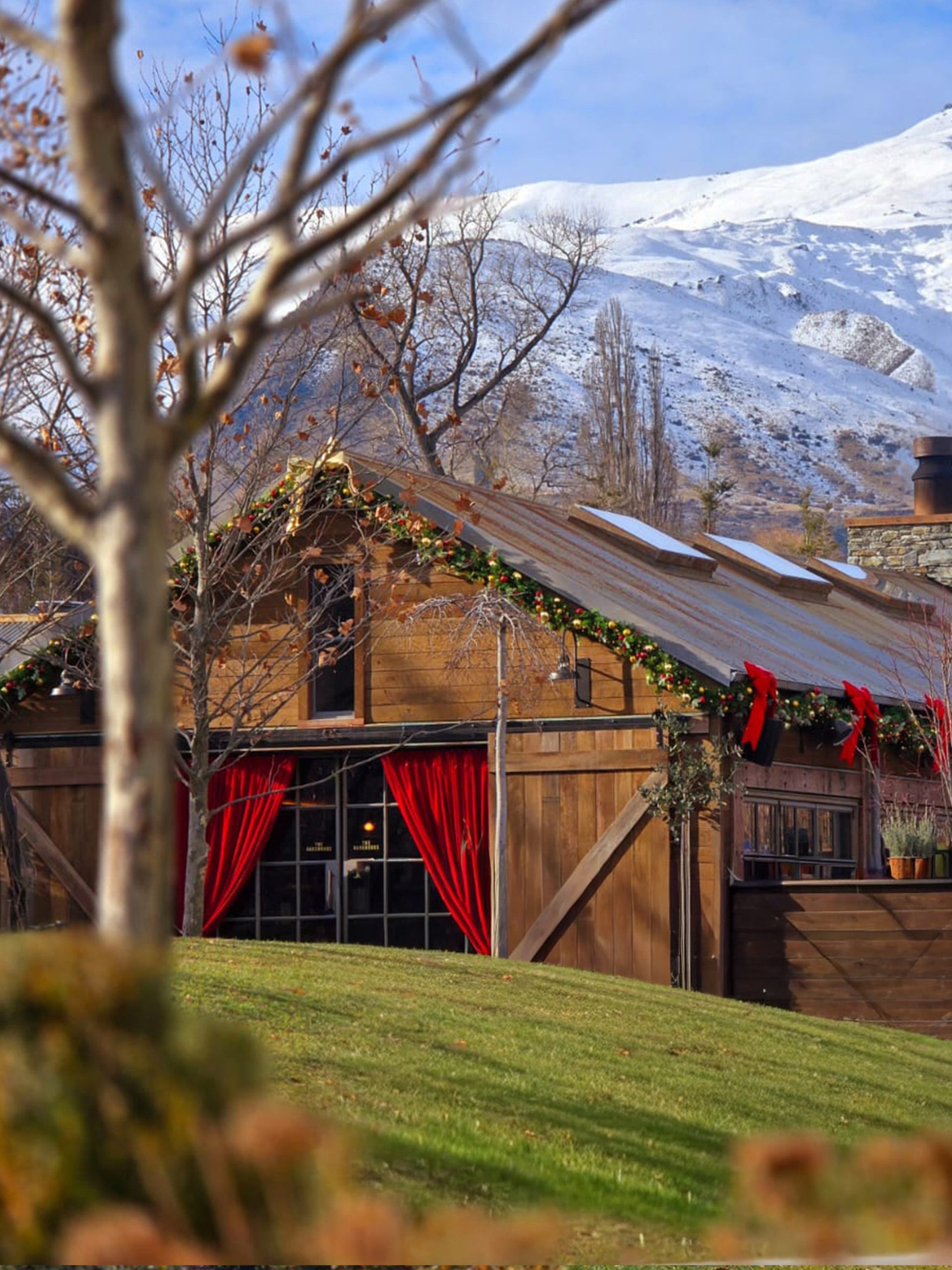 Christmas-decorated rustic wooden barn with red curtains and garlands against a snow-capped mountain.