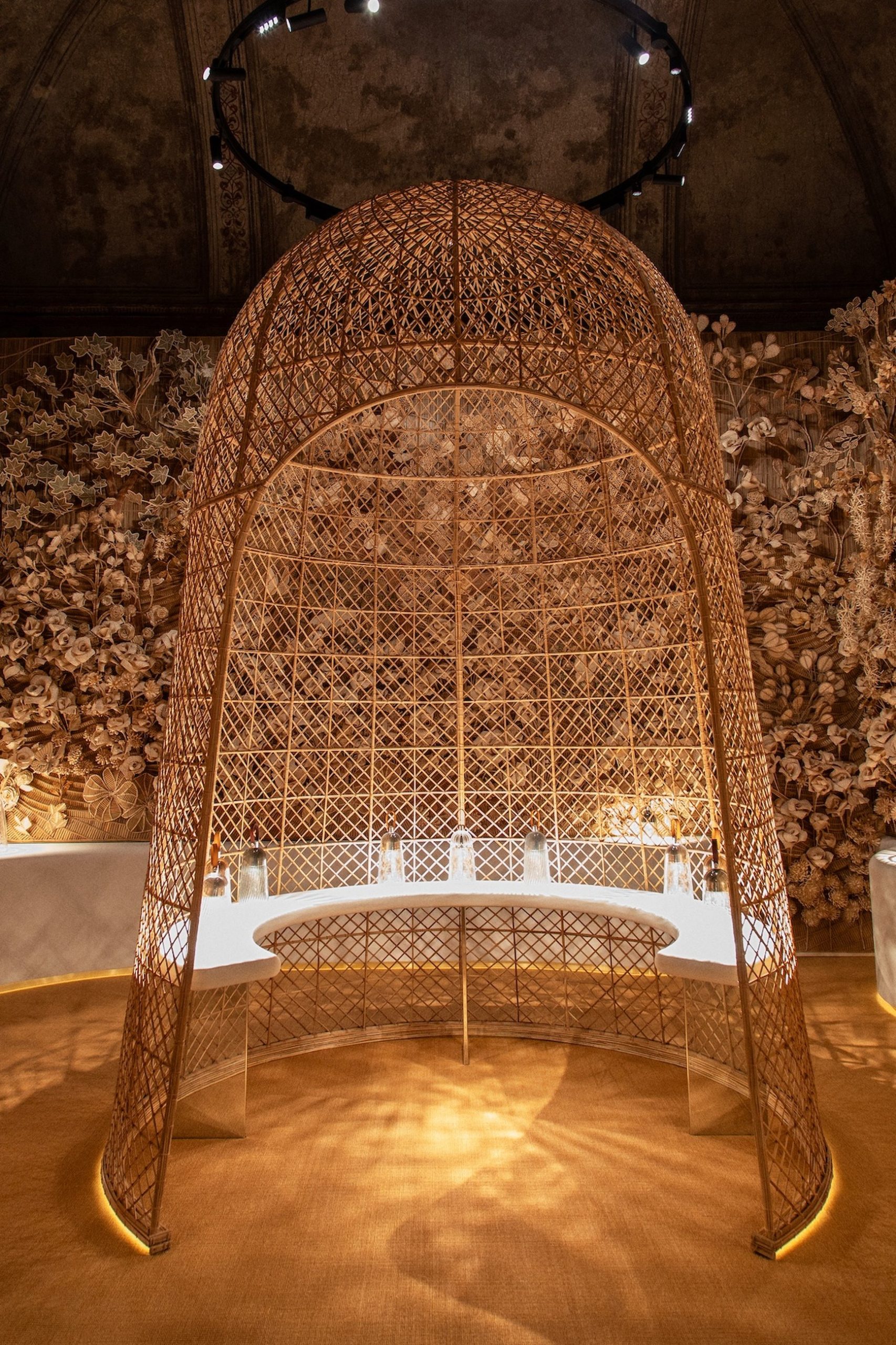Large domed woven rattan lattice structure enclosing a circular dining table in a floral-adorned interior.
