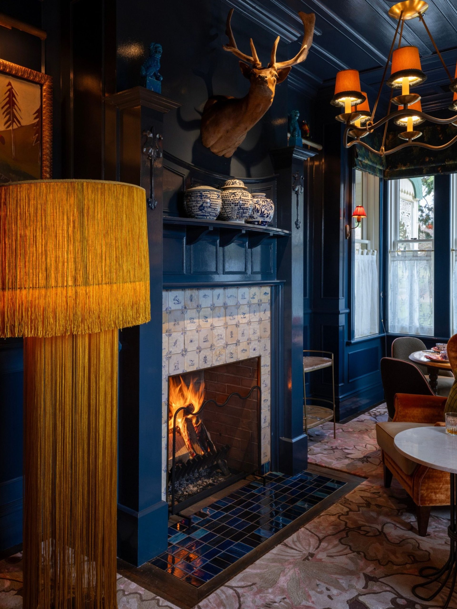 Dark navy lounge with roaring fireplace, stag taxidermy, Delft tiles, velvet chairs and gold fringed lamp.