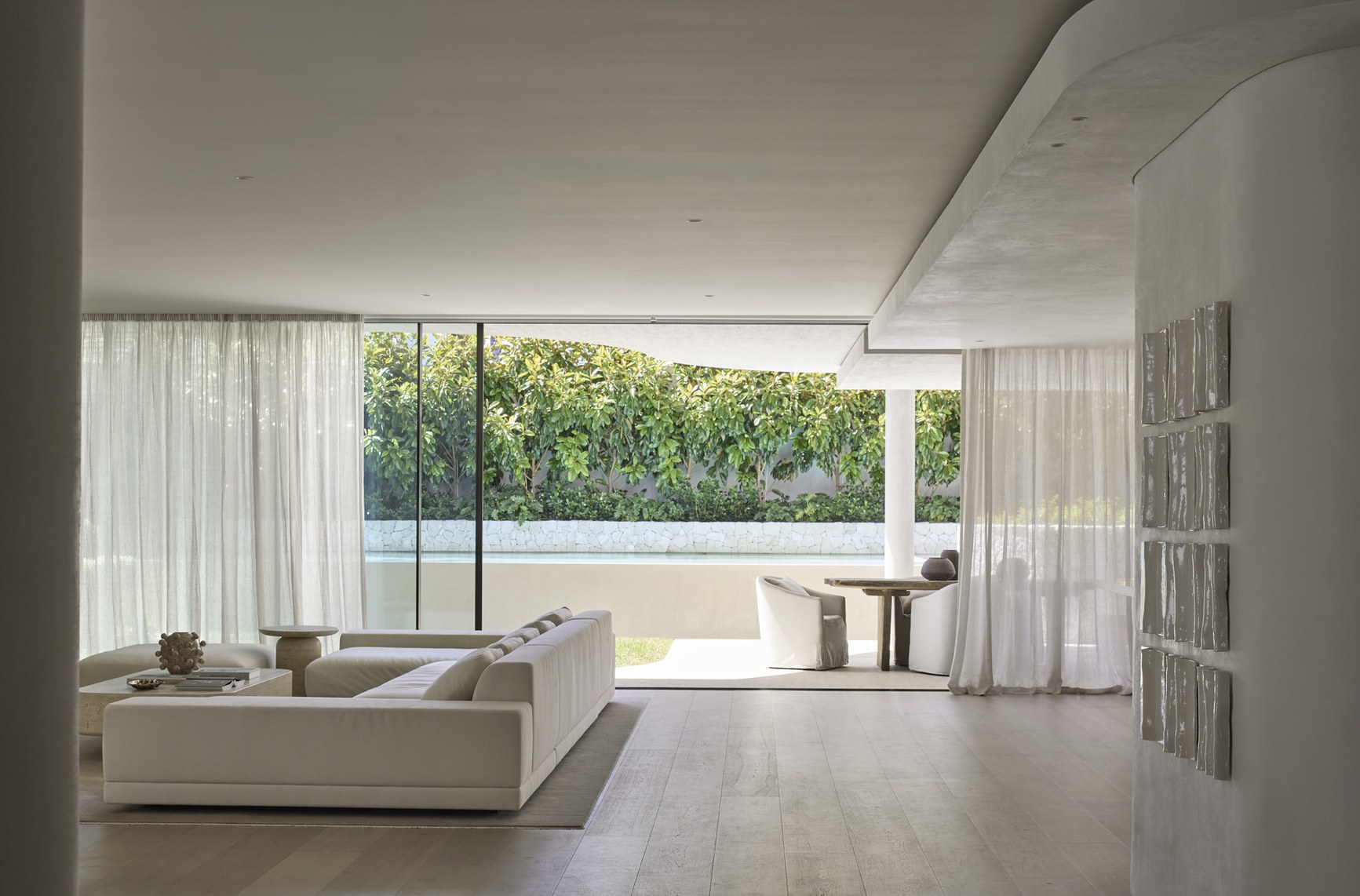 Minimalist white open-plan living room opening to a lush garden and pool terrace.
