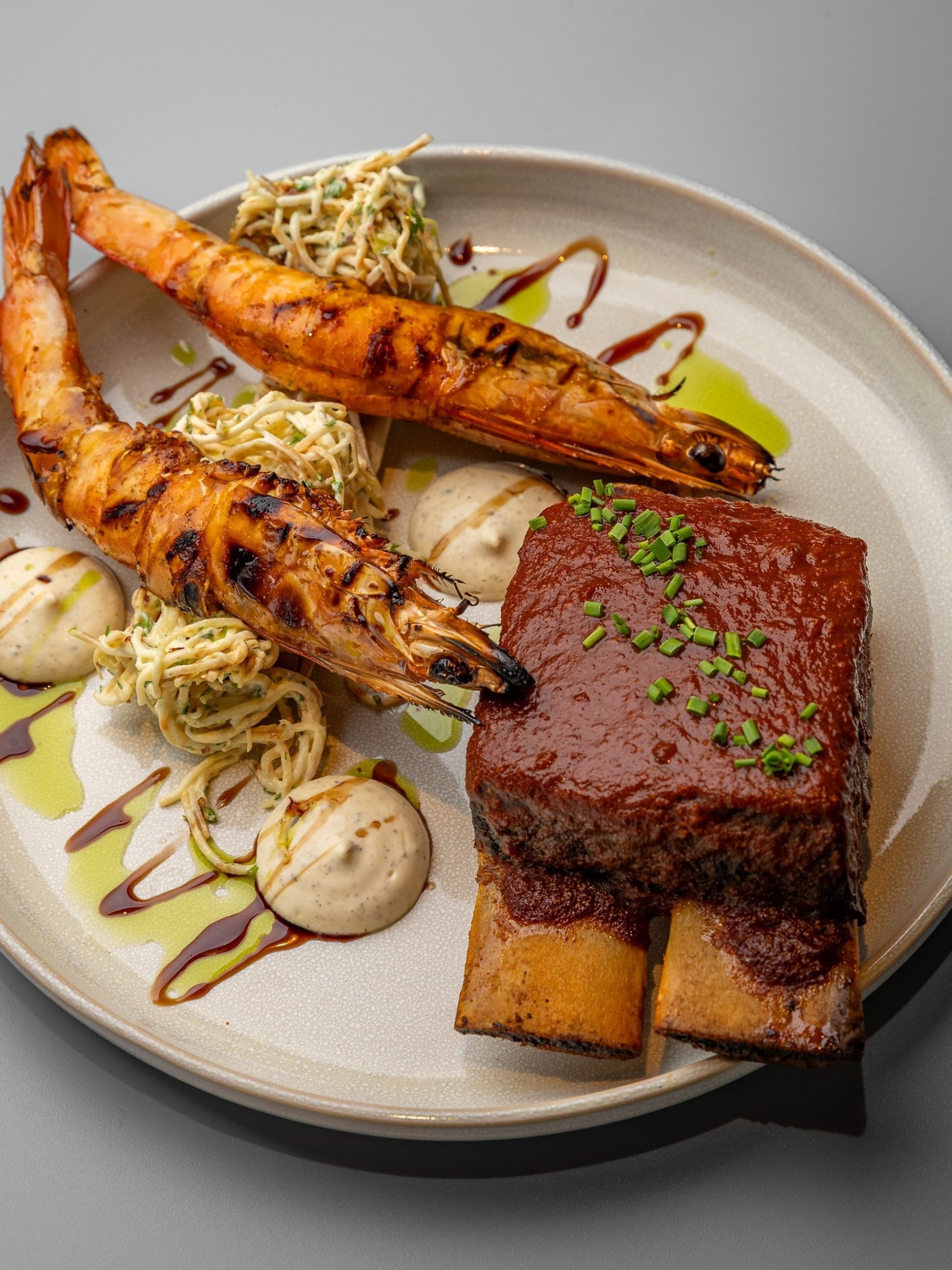 Grilled tiger prawns and glazed beef short rib with sesame noodles on a ceramic plate.