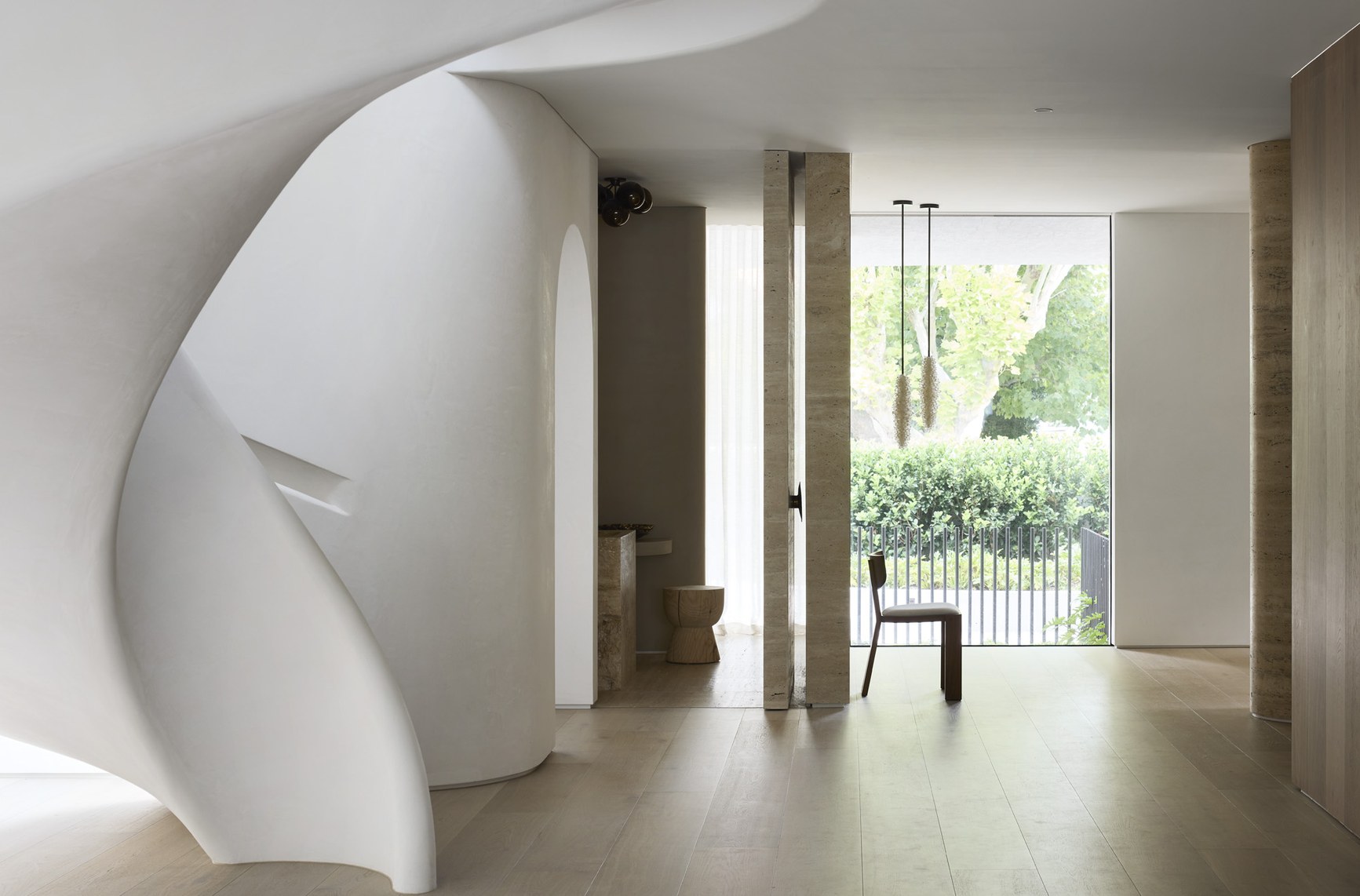 Curved white sculptural staircase in a minimal luxury interior with travertine columns and garden views.