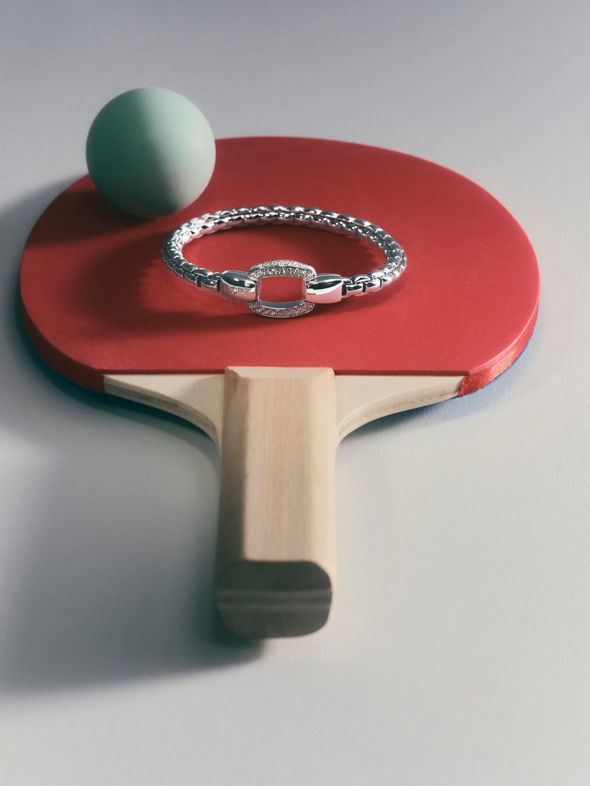 Silver diamond bracelet resting on a red table tennis paddle with a green ping-pong ball.