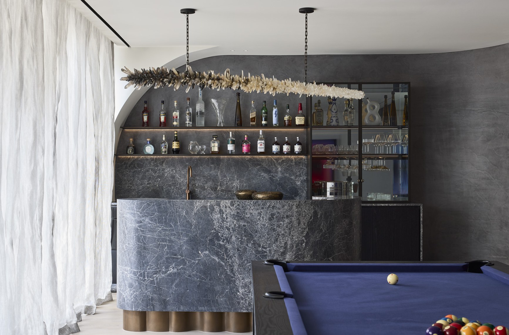 Luxury home bar with dark marble counter, crystal chandelier, spirits shelving, and blue pool table.