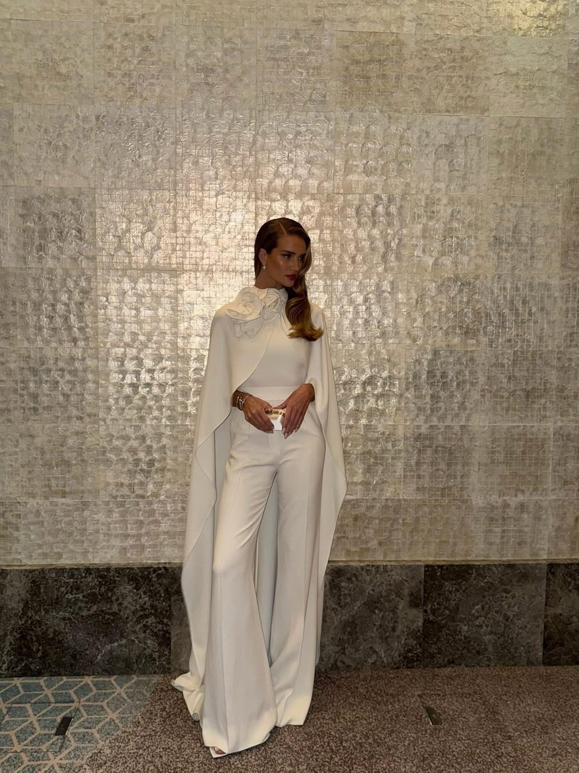 Woman in white caped wide-leg jumpsuit with floral neckline posing against silver textured wall.