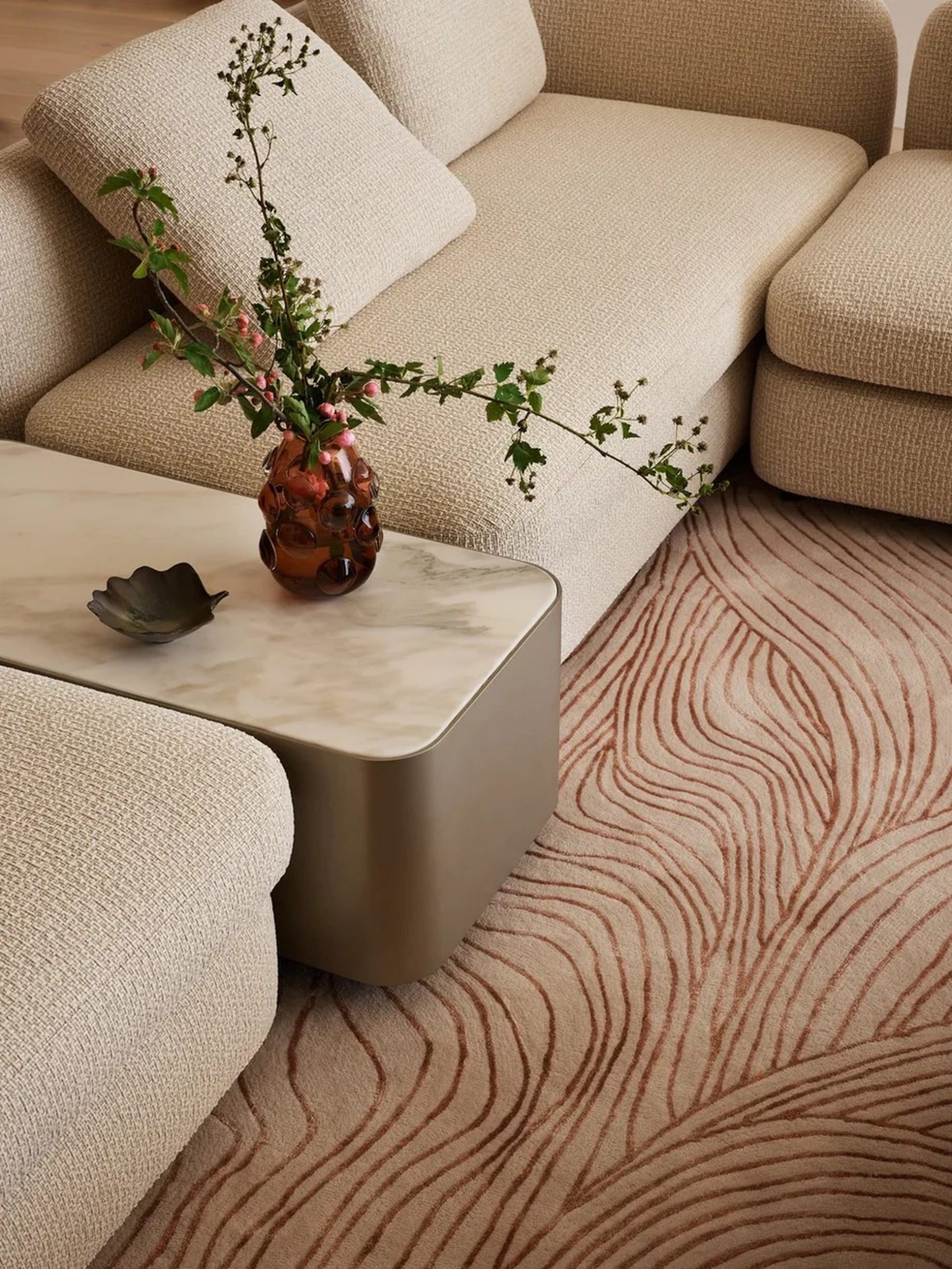 Bouclé sectional sofa with marble coffee table, amber vase, and wave-patterned rug in warm neutrals.