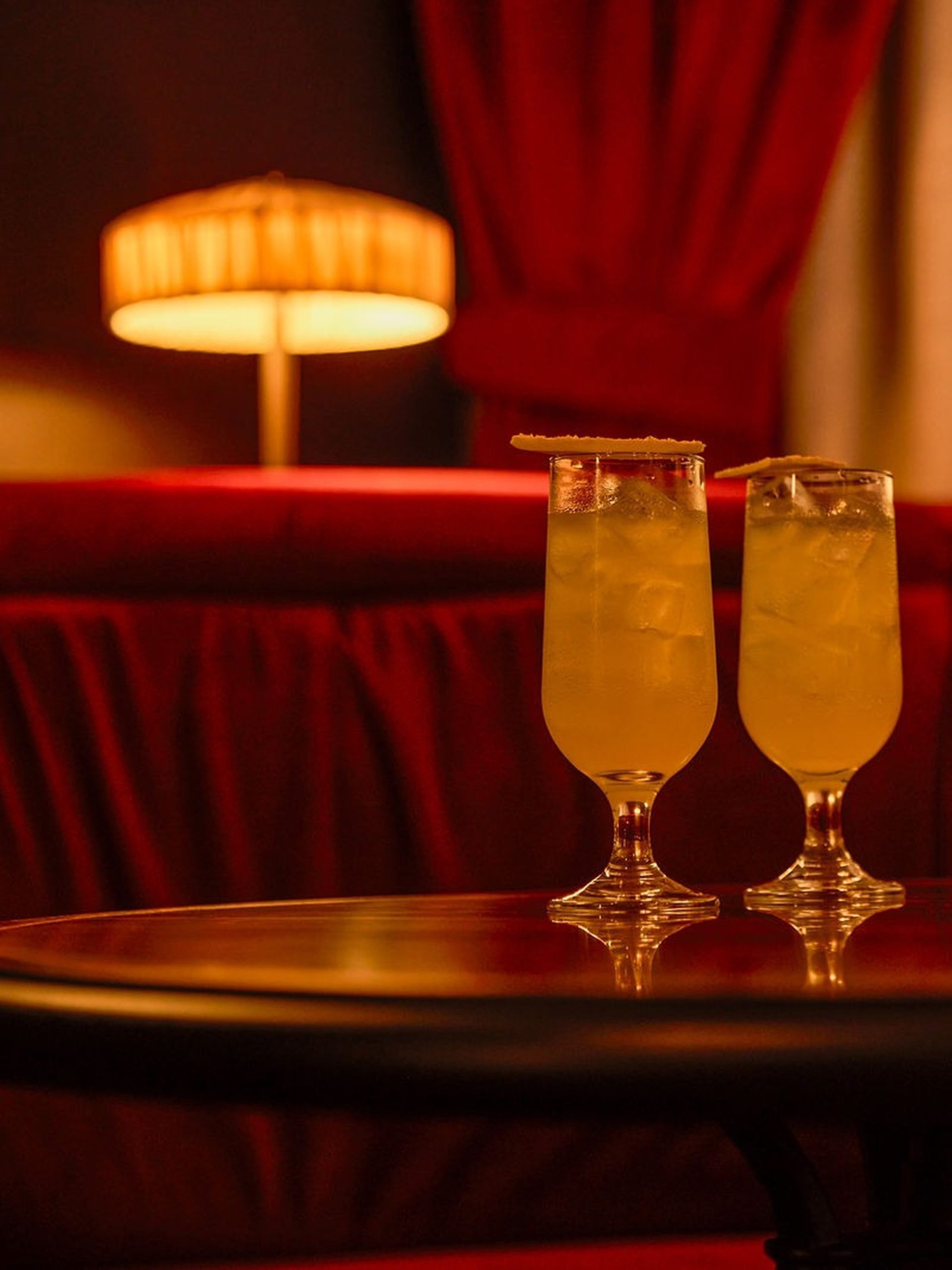 Two amber cocktails in pilsner glasses on a dark table inside a red velvet bar.