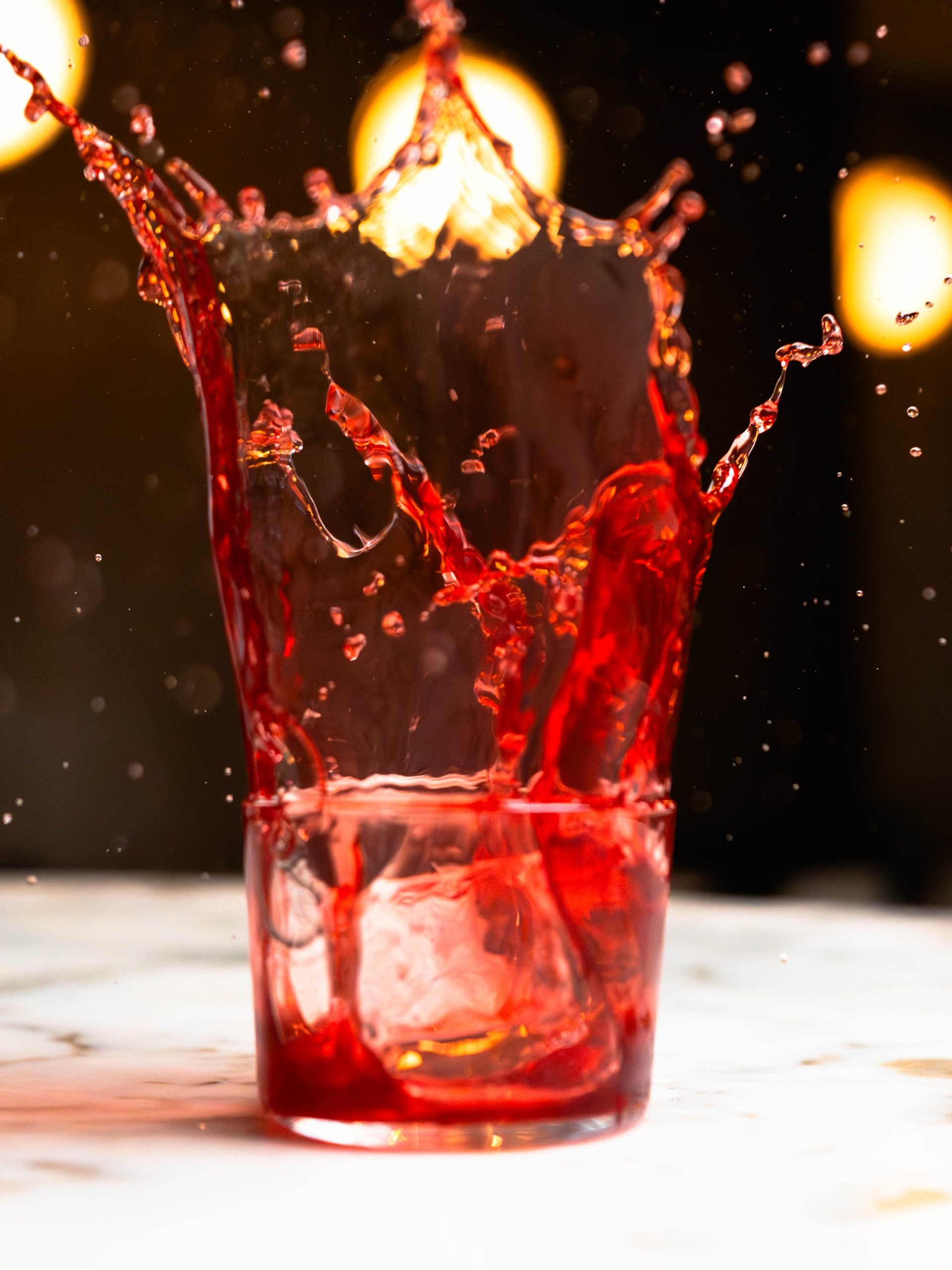 Crown-shaped red liquid splash bursting from an ice-filled rocks glass on a marble surface.