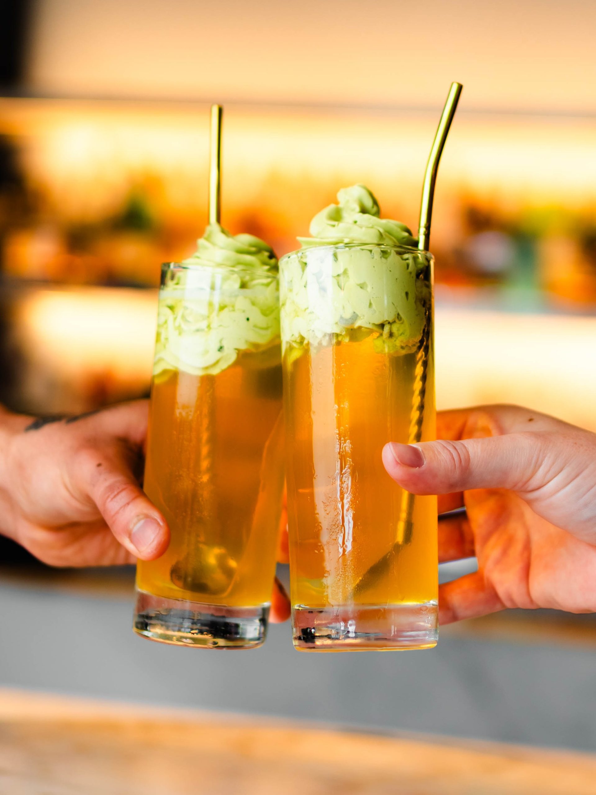 Two hands toasting with tall golden mocktail floats topped with matcha soft-serve and gold straws.