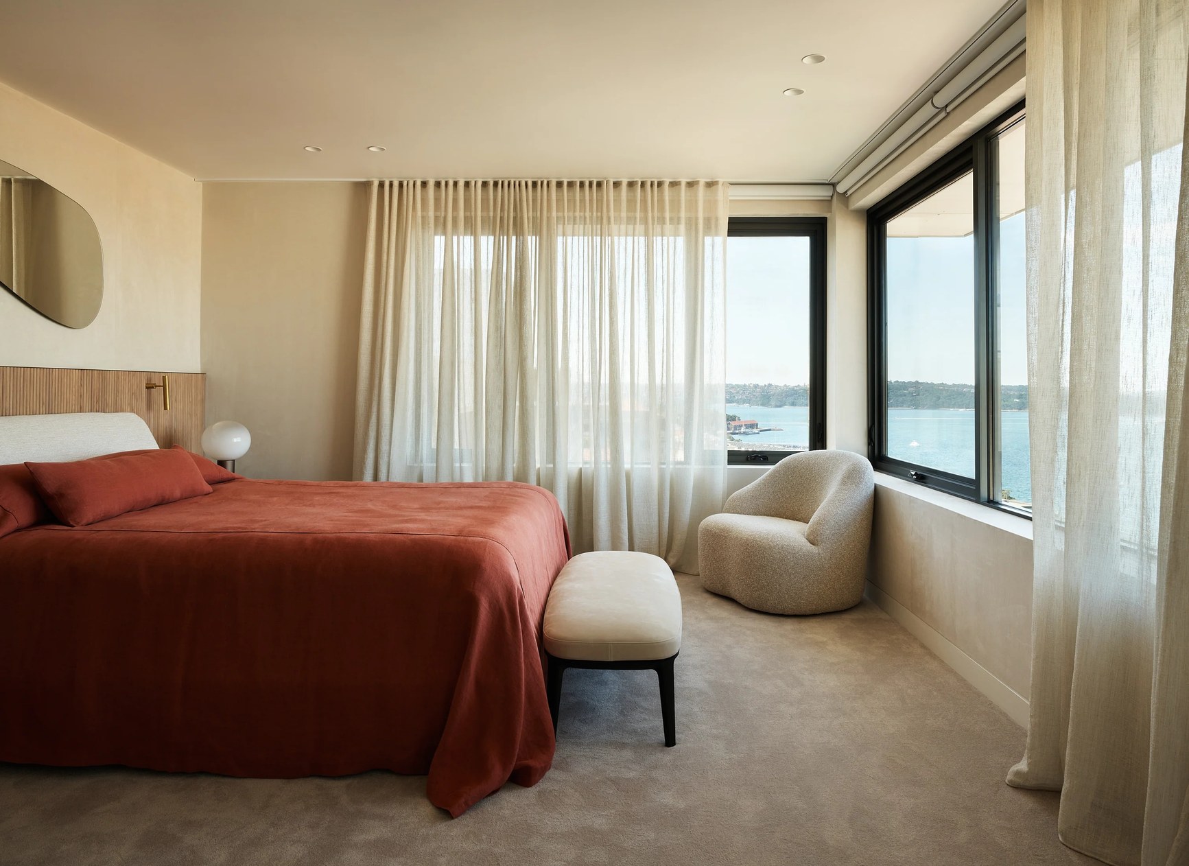 Luxury bedroom with terracotta bedding, bouclé armchair, and panoramic harbour views through large windows.