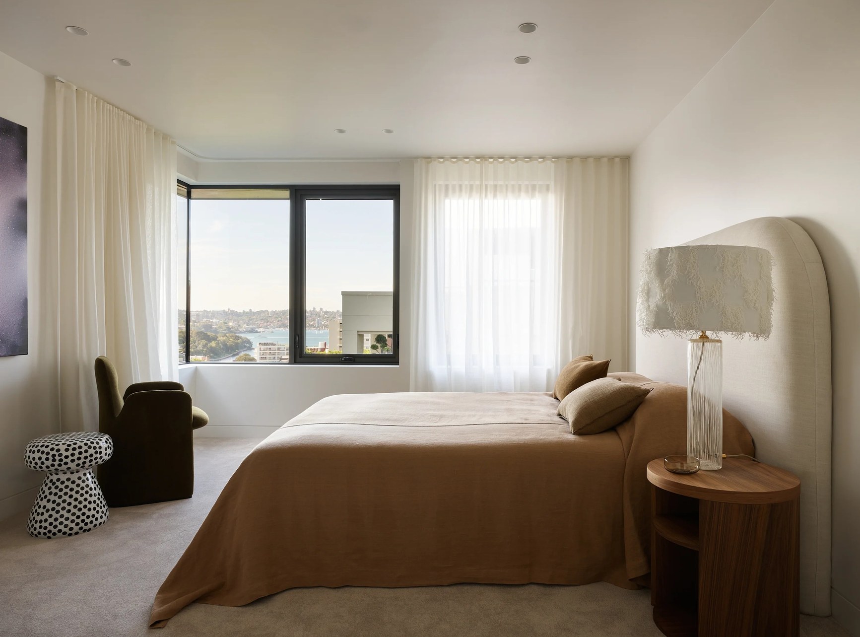 Contemporary bedroom with caramel bedding, arched headboard, sheer curtains and sweeping harbour views.