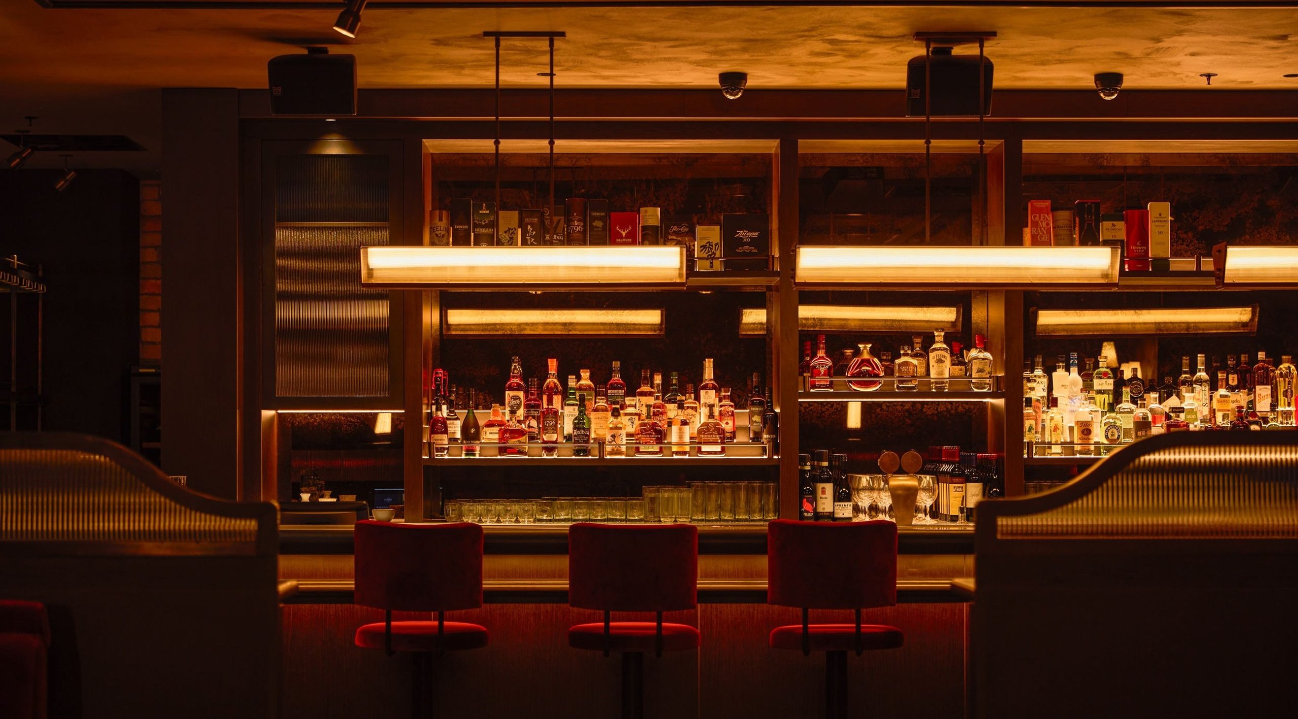 Empty upscale cocktail bar with red velvet stools and warmly lit shelves of premium spirits.